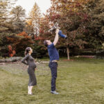 dad throwing toddler son into the air while pregnant mother looks on adoringly during family portraits in James Gardens