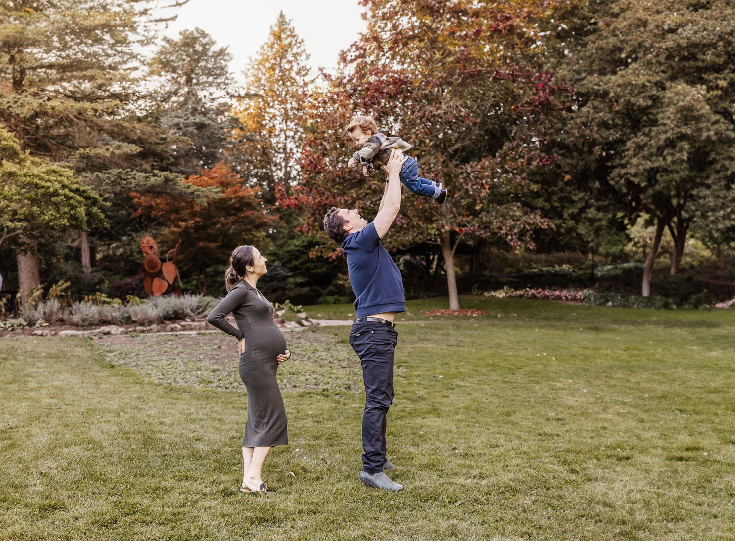 dad throwing toddler son into the air while pregnant mother looks on adoringly during family portraits in James Gardens