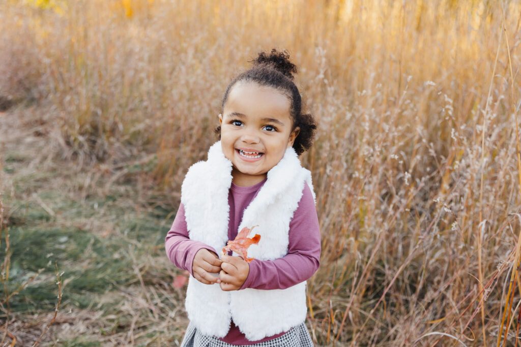 toddler girl giggling in golden tall grass. She is holding a leaf.