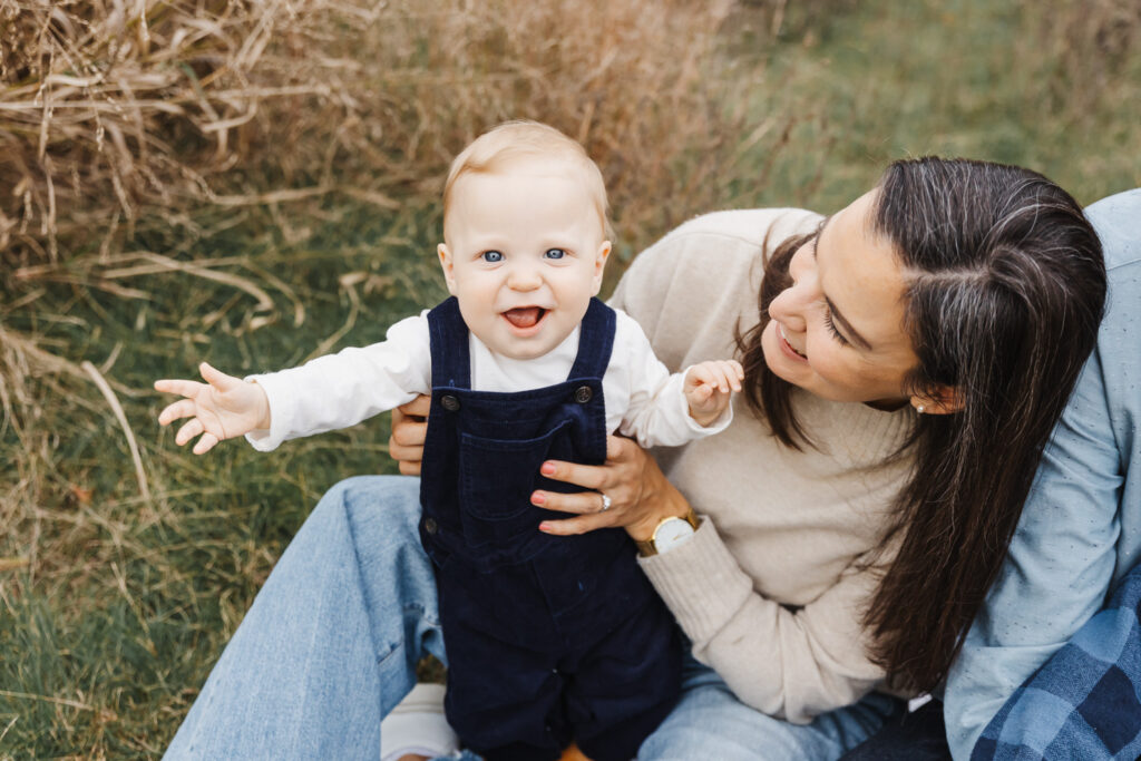 mom holding baby boy in standing position. He is smiling at the camera while his mom smiles at him.