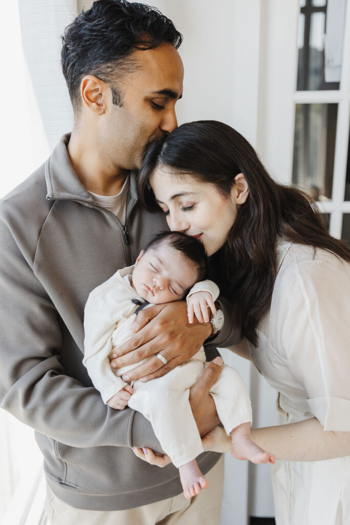 man tenderly kisses woman as she kisses her newborn baby's head.