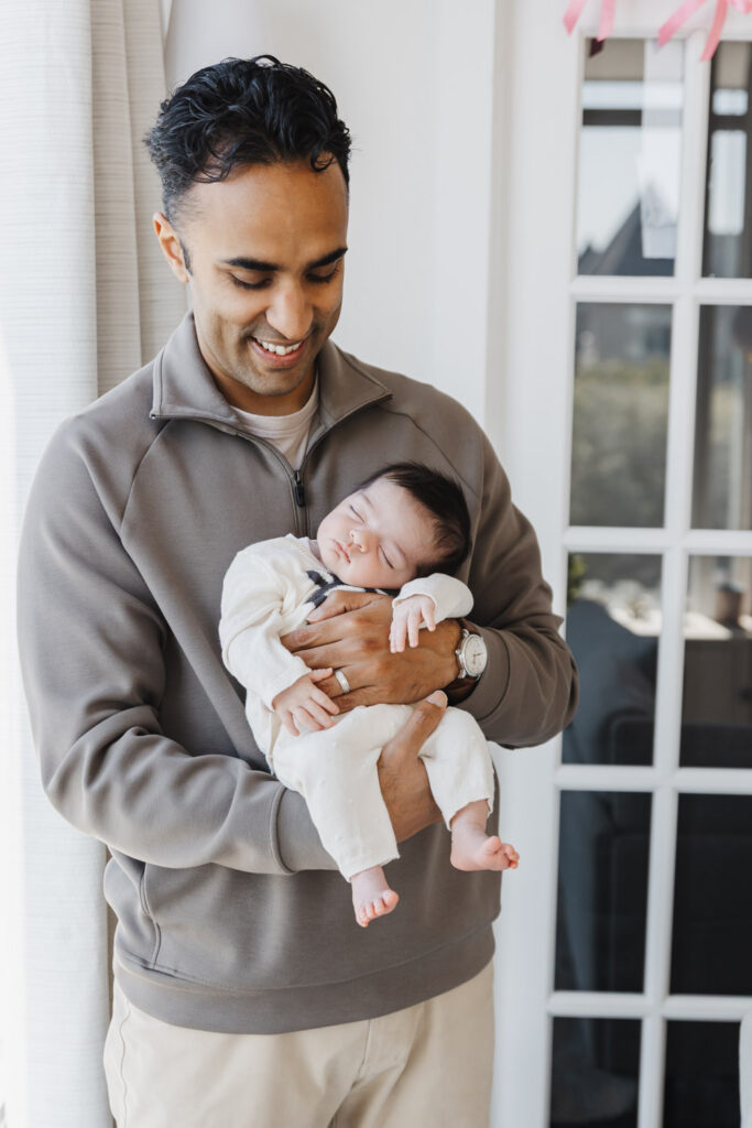 Dad dressed in brown sweater holding newborn daughter dressed in white knit outfit