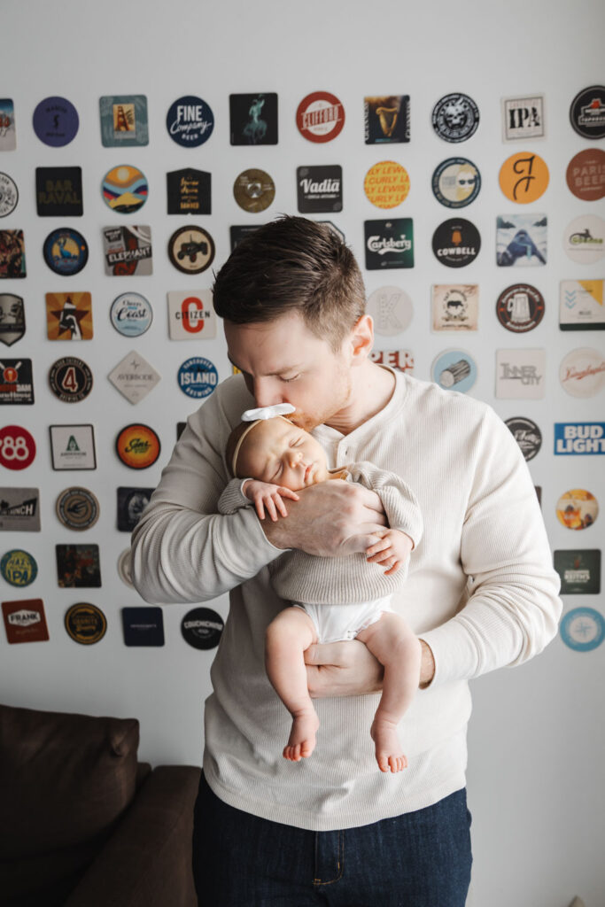 Dad dressed in comfy oatmeal henley shirt, kissing newborn daughter